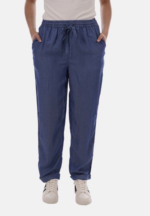 Personne portant un pantalon bleu marine ample avec cordon de serrage et poches, ainsi que des baskets blanches à rayures noires, debout devant un fond uni.