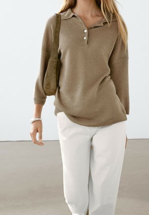 Vrouw draagt een bruin gebreide polotrui, witte broek, een bruine schoudertas en een zilveren armband, staand tegen een neutrale achtergrond.