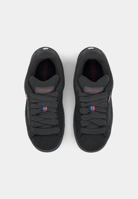 Black sneakers with a textured upper, flat laces, and a red and blue accent near the tongue. Featuring a round logo patch on the side.