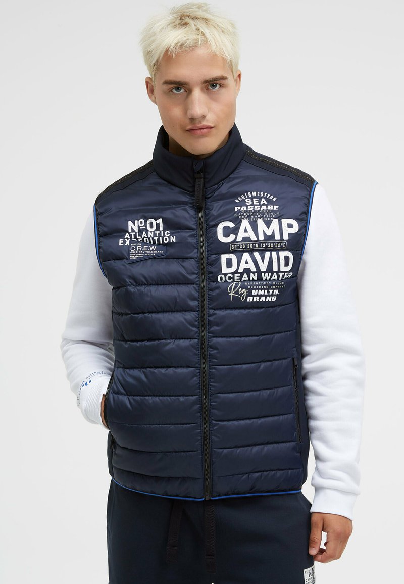 Marinefarbener gepolsterter Weste mit hohem Kragen, Reißverschluss und weißen Textelementen. Verfügt über horizontale Steppnähte und Seitentaschen.