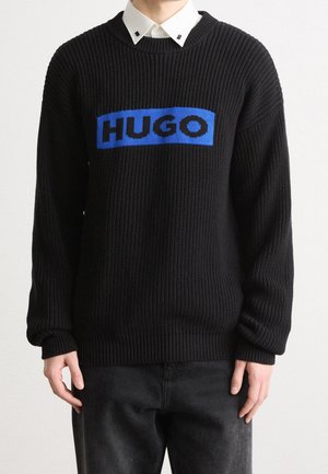 Osoba nosící černý žebrovaný svetr s modrou obdélníkovou náplastí a černým nápisem "HUGO", vrstvený přes bílou košili s límečkem.