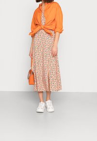 Blumiges Midi-Kleid mit einem hellen Pfirsich-Hintergrund, orangefarbenem gebundenem Shirt, weißen Sneakers und einer orangefarbenen Handtasche mit einer Kettenriemen.