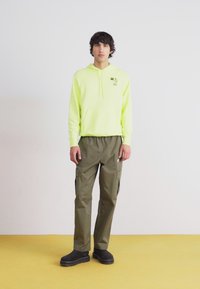 Lys gul hoodie med kængurulomme, olivengrøn cargoset bukser og sorte slip-on sko, mod en lys baggrund og gul gulv.