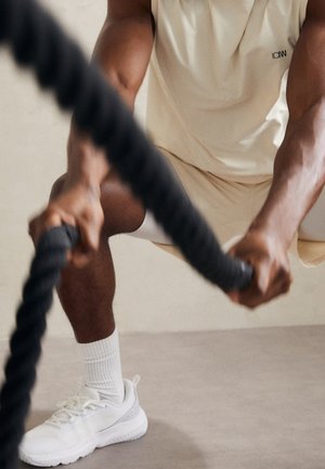 Sportowiec w beżowych szortach i koszulce bez rękawów wykonuje w pomieszczeniu ćwiczenie z liną do walki, trzymając grubą czarną linę, nosząc białe buty sportowe i skarpetki.