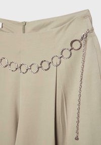 Jupe beige avec une ceinture chaîne argentée composée de maillons circulaires texturés et une chaîne de fermeture prolongée pendante sur le côté.