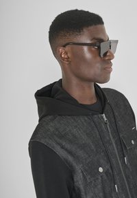 Chaqueta con capucha de mezclilla negra con herrajes plateados y dos bolsillos en el pecho, llevada sobre una camiseta de manga larga negra; modelo con gafas de sol oscuras.