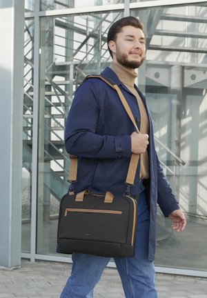 Man in navy coat and tan turtleneck carries black and tan shoulder bag walking outdoors in front of glass building with stairs.