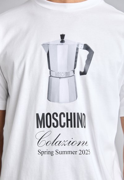 T-shirt blanche présentant un graphisme d'une cafetière moka métallique avec une poignée noire, et le texte "MOSCHINO Colazione Printemps Été 2025" en dessous.