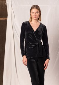 Svart långärmad omlottopp i sammet med V-ringning och knutdetalj framtill. Mjuk textur, figurnära passform och matchas med svarta byxor i samma material.