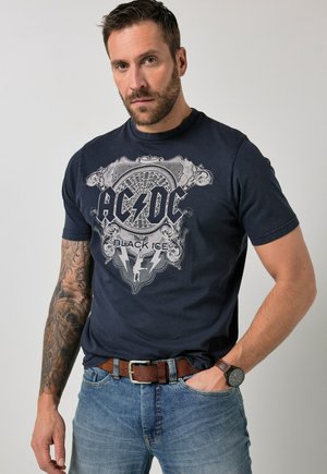 Mężczyzna z brodą, ubrany w czarną koszulkę z grafiką AC/DC Black Ice, niebieskie dżinsy, brązowy pasek, zegarek na nadgarstku oraz rękaw z tatuażami na lewej ręce.