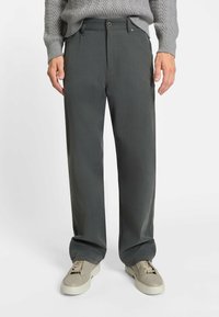 Dunkelgraue, weit geschnittene Hosen mit glatter Textur, Gürtelschlaufen, Knopfverschluss und subtilen Steppdetails, kombiniert mit hellen Sneakers.