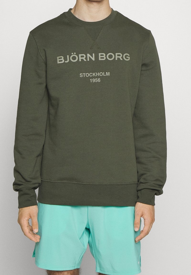 Homme portant un sweat-shirt vert olive avec le texte "BJÖRN BORG STOCKHOLM 1956" et un short turquoise clair, debout devant un fond uni.
