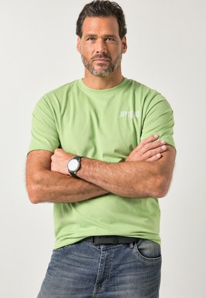 Hellgrünes Baumwoll-T-Shirt mit kurzen Ärmeln und kleinem weißem "JP1880"-Logo. Kombiniert mit blauen Jeans und einer schwarzen Uhr.