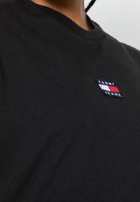 T-shirt en coton noir avec un col rond, arborant un logo rectangulaire brodé en rouge, blanc et bleu sur la poitrine gauche.