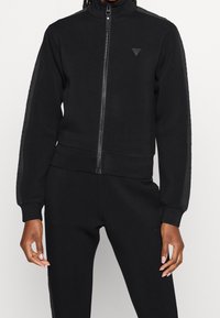 Person trägt schwarze Jacke mit Reißverschluss und passende schwarze Hose mit dezenten Logo-Aufnähern auf Brust und Oberschenkel, steht vor schlichem Hintergrund.