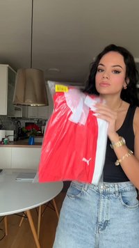Jauna sieviete ar tumšiem matiem un zelta rotaslietām tur sarkanu Puma sporta krekliņu plastmasas iepakojumā mūsdienīgā virtuvē.