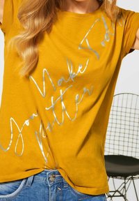 Hořčicově žluté tričko s krátkým rukávem, na kterém je stříbrný nápis "Děláš dnešek lepší." Měkká bavlněná textura, uvolněný střih.