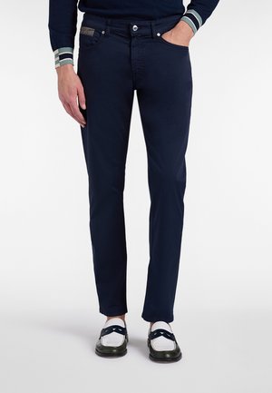 Mand iført mørkeblå slanke bukser, sort-hvide loafers og en mørk sweater med stribede manchetter, stående med den ene hånd i lommen.