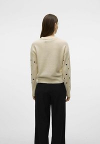 Pull crème avec des motifs de cœurs noirs sur les manches, bord-côtes aux poignets et à l'ourlet. Assorti avec un pantalon large noir. Texture tricotée, unie et douce.