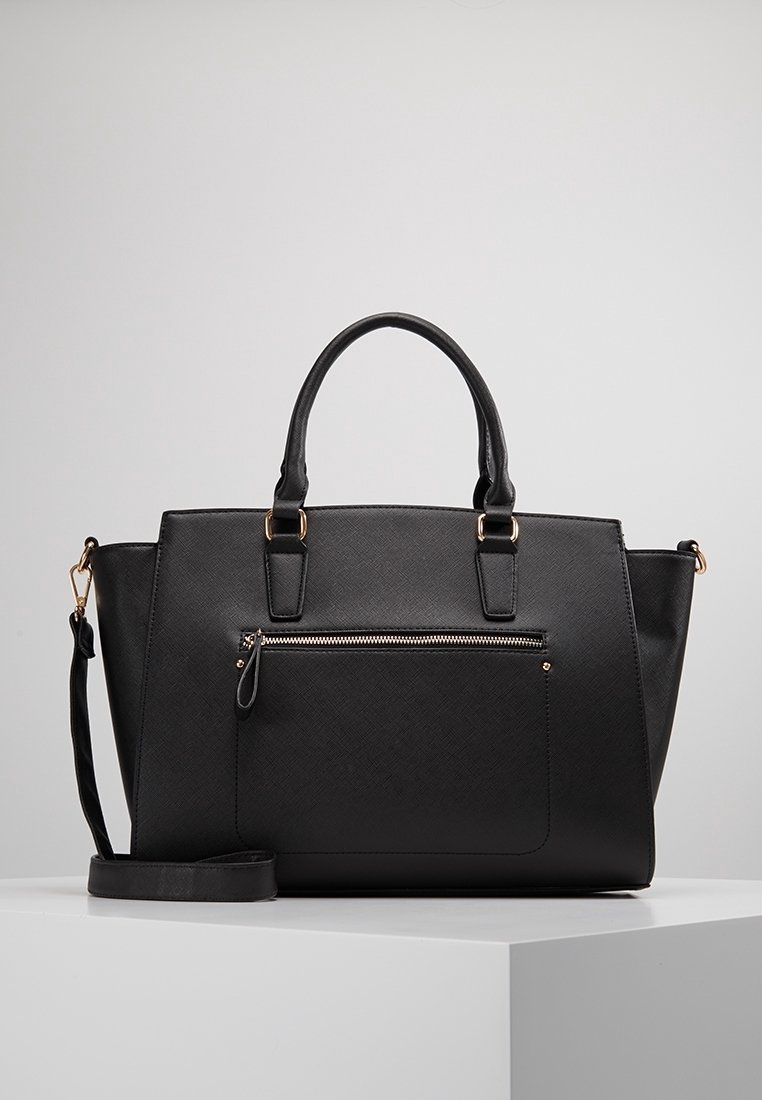 Sac cabas en cuir noir texturé avec double poignée, poche zippée avant et bandoulière amovible, présenté sur un piedestal blanc.