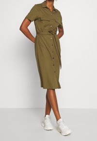 Robe boutonnée vert olive à manches courtes, deux poches poitrine, taille ceinturée et ourlet mi-long, portée avec des baskets blanches.