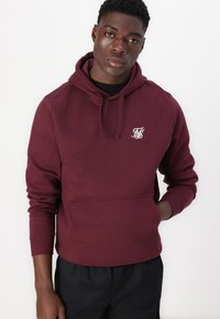 Hoodie bordeaux en mélange de coton avec une poche avant, un capuchon à cordon de serrage et un petit logo blanc sur la poitrine. Présente des poignets côtelés.