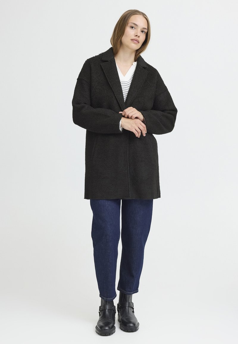 Cappotto in misto lana nero, strutturato, con colletto a scialle, maniche lunghe e tasche frontali, abbinato a jeans blu scuri alla caviglia e stivaletti neri.