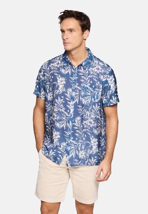 Hombre con camisa azul de manga corta con estampado floral y pantalones cortos beige, de pie con las manos en los bolsillos contra un fondo liso.