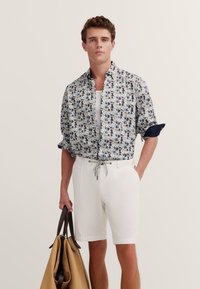 Kurzarmhemd mit blauem und floralem Muster, kombiniert mit weißen Shorts. Hält eine braune Handtasche mit dunkelbraunen Akzenten.
