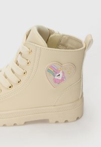 Zapatilla alta beige de material sintético, con ojales dorados, suela texturizada y un parche en forma de corazón con un diseño de unicornio.