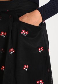 Gonna nera con tessuto a rilievo e ricami floreali rossi, tasche sui lati per funzionalità e una superficie testurizzata.
