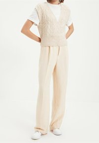 Maglioncino beige senza maniche con scollatura a V e motivo a rombi, abbinato a pantaloni larghi beige chiaro e sneakers bianche.