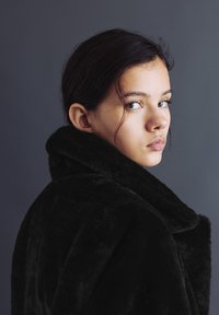 Tânără cu păr închis la culoare, privind peste umăr, purtând o haină neagră pufoasă, pe un fundal gri simplu.