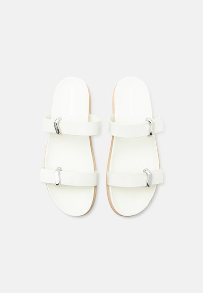 Άσπρα επίπεδα σανδάλια με δύο φαρδιές λουριά το καθένα, με ασημένιες αγκράφες, απεικονιζόμενα από πάνω σε ανοιχτό γκρι φόντο.