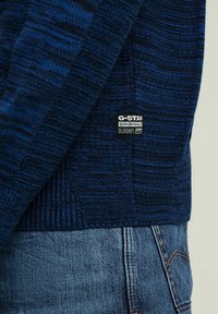Maglione lavorato a maglia blu scuro con un motivo testurizzato, polsini a coste e un'etichetta G-Star Originals sul lato. Abbinato a jeans in denim chiaro.