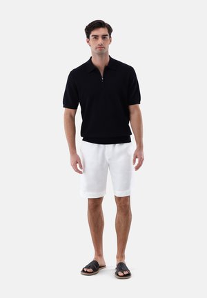Homme portant un t-shirt noir à manches courtes avec col zippé, un short blanc jusqu'aux genoux et des sandales noires, debout sur un fond uni.