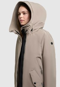 Beige hooded jacket with a quilted interior, black zipper, side and snap-button pockets, and smooth texture, designed for versatility and warmth.