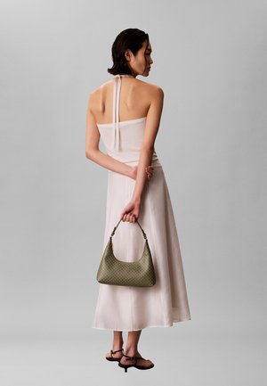 Naine heleroosa kaelusega kleidis, hoides selja taga rohelist mustrilist käekotti, seljas mustad madala kontsaga sandaalid, seismas halli tausta ees.