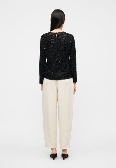 Μαύρο, υφασμάτινο πλεκτό τοπ με μακριά μανίκια και άνοιγμα πίσω στο κέντρο, συνδυασμένο με φαρδιά μπεζ παντελόνια και μαύρα παπούτσια με τακούνι.