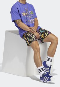 Maglietta grafica viola con design colorati, pantaloncini neri a fantasia e scarpe da ginnastica blu con calzini a righe bianche e nere.