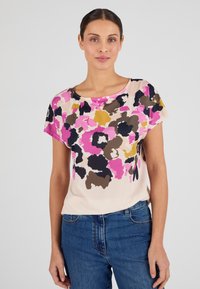 Mujer con una blusa de manga corta con estampado floral abstracto en tonos rosa, negro, marrón y mostaza, combinada con jeans azules, frente a un fondo liso.