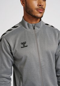 Veste zippée grise en tissu lisse et texturé. Présente un logo noir et des accents d'épaule avec un motif en chevrons. Fermeture éclair à l'avant.
