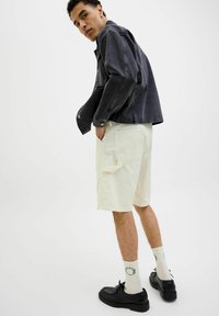 Veste en denim noir, shorts cargo crème avec poches latérales, chaussures noires à lacets et chaussettes blanches avec accents imprimés. Style simple et contemporain.