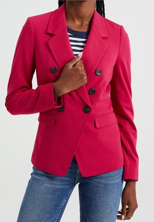 Woman wearing a bright pink double-breasted blazer over a navy striped shirt and blue jeans, holding the blazer lapel.
