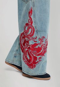 Light blue denim jeans with wide legs featuring a large red embroidered dragon design on one leg above black shoes.