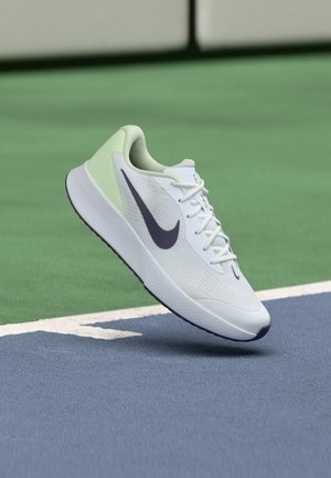 Witte en lichtgroene Nike hardloopschoen met donker logo zwevend boven een blauwe en groene tennisbaan.