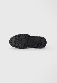 Suela de zapato de goma negra con surcos y hendiduras profundas para mayor tracción. Presenta un acabado suave y una marca en relieve en la parte inferior.