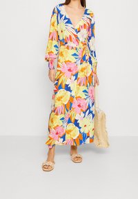 Vestido envolvente de flores con colores vibrantes, mangas largas con puños elásticos y escote en V. Acompañado de un bolso de palma y sandalias.