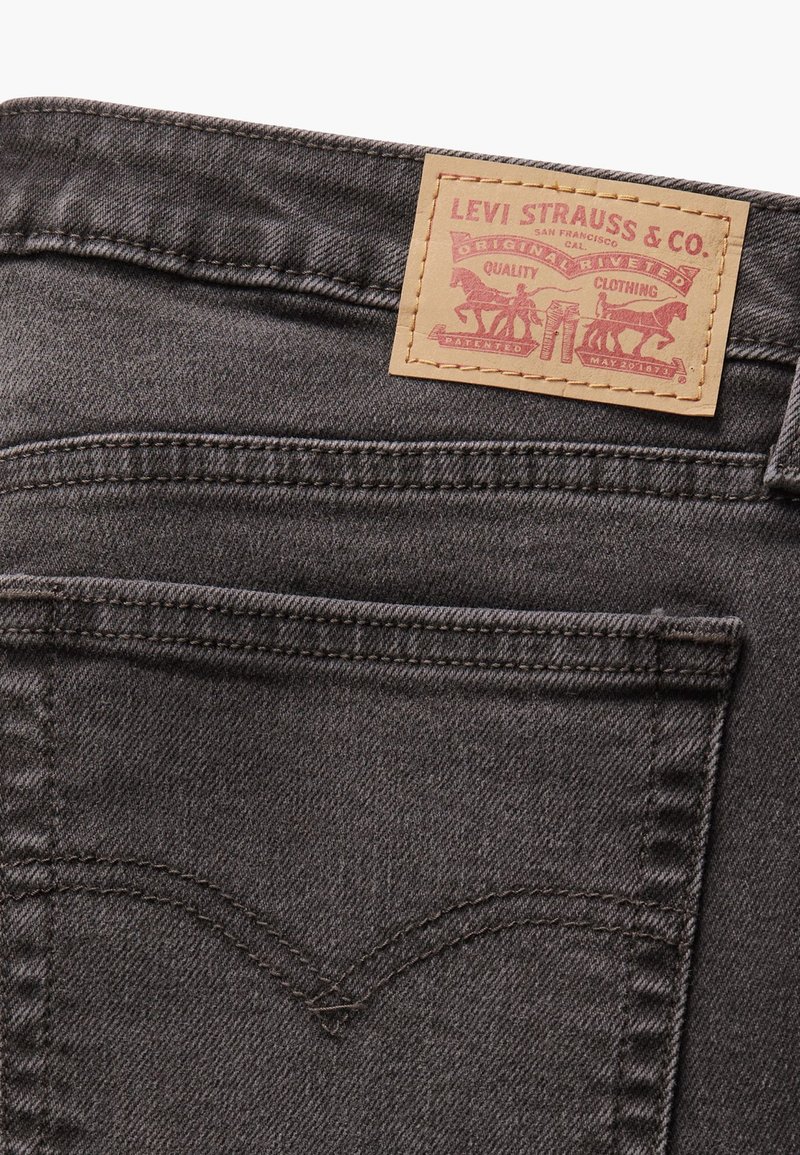 Jeans en denim gris avec une patch en cuir tan rectangulaire arborant un texte rouge et deux graphiques de chevaux, mettant en avant un design classique et des détails de couture.