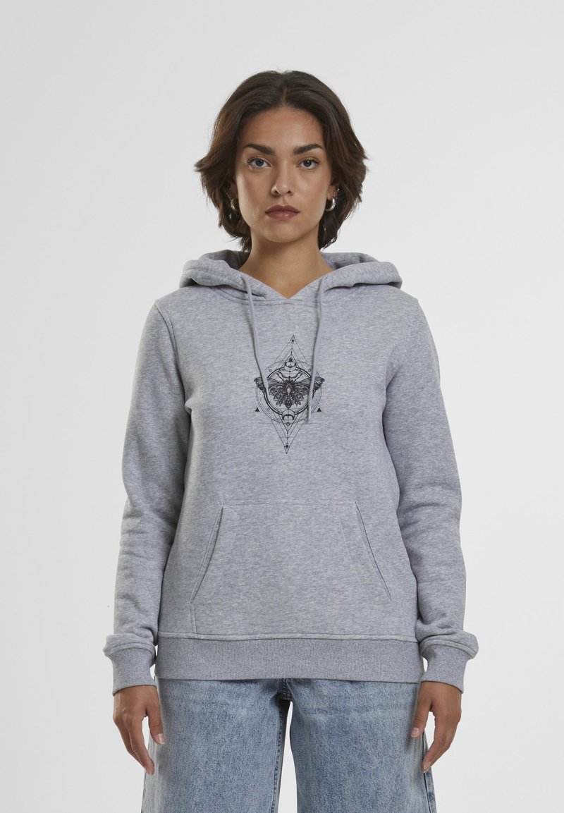 Femme portant un sweat à capuche gris clair avec un motif géométrique noir d'insecte, debout devant un fond clair uni.